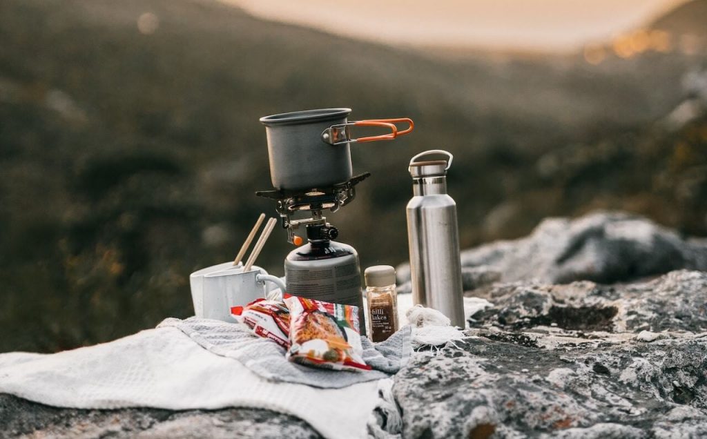 Ein Bild von einem Campingkocher mit einer kleinen Pfanne darauf und einer Wasserflasche daneben, der auf einem Felsen mit majestätischen Bergen im Hintergrund aufgestellt ist.