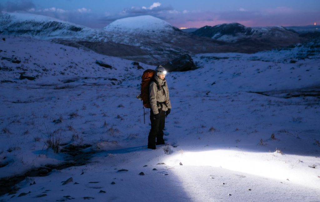 Ein Bild von einem Wanderer, der durch die verschneite Nacht wandert und eine Stirnlampe trägt, um den Weg zu beleuchten.